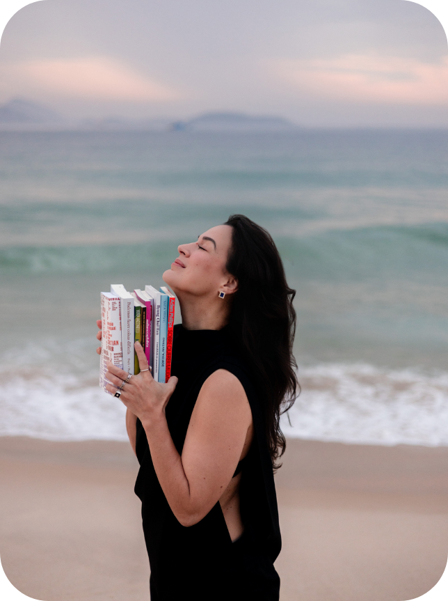 Karina Francis de olhos fechados segurando livros junto ao peito, à beira-mar