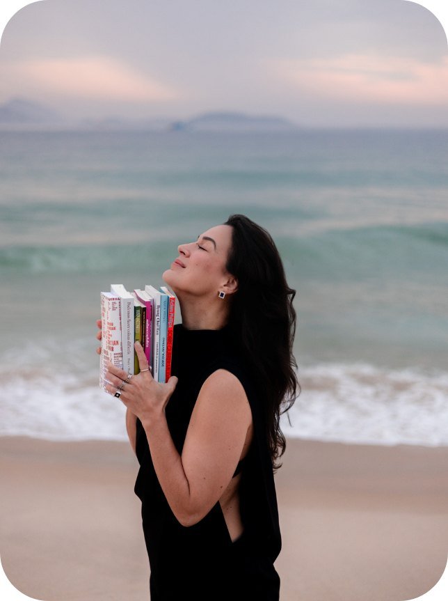 Karina Francis de olhos fechados segurando livros junto ao peito, à beira-mar